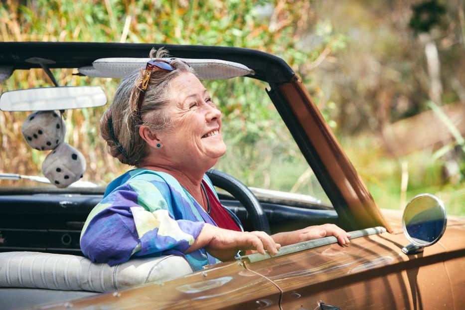 Eine ältere Frau lächelt, während sie in einem braunen Cabriolet fährt.