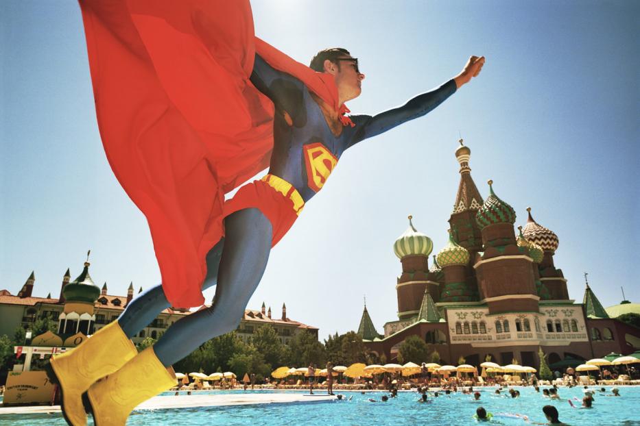 Ein Mann im Superman-Kostüm springt vor einem Schwimmbad mit russisch anmutender Architektur.
