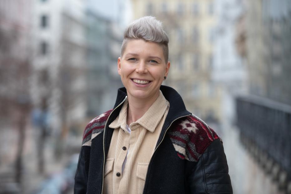 Eine lächelnde Frau mit kurzen, grauen Haaren und einer gemusterten Jacke steht im Freien.