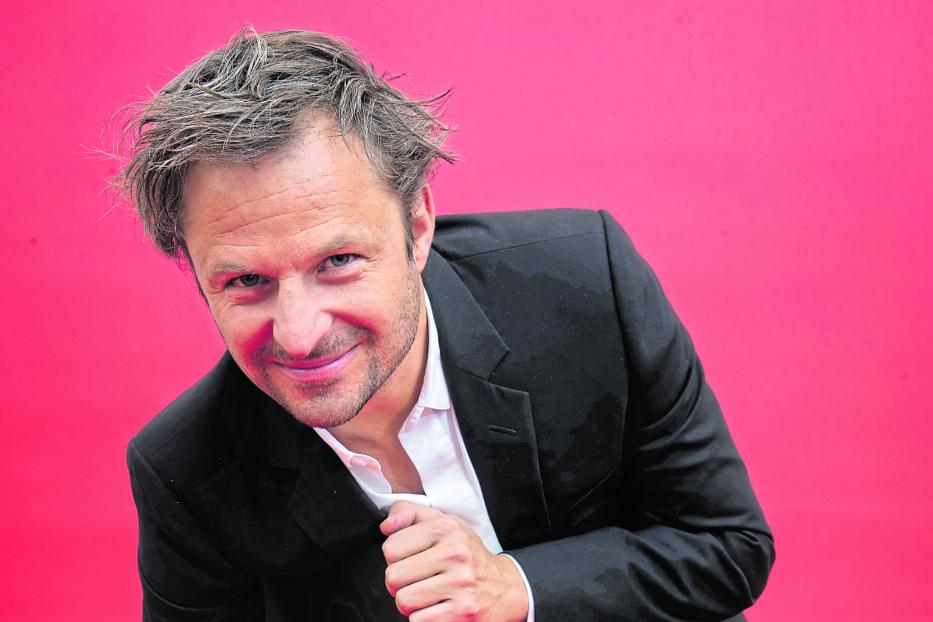 Philipp Hochmair sitzt auf einem Stuhl vor einem roten Hintergrund.