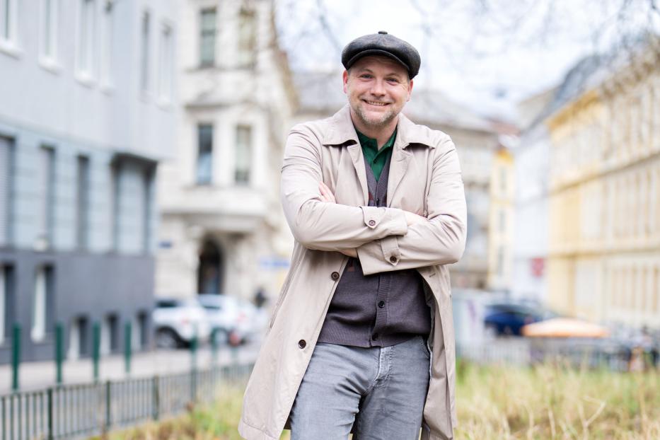 Ein lächelnder Mann mit Schirmmütze und Trenchcoat steht auf der Straße.