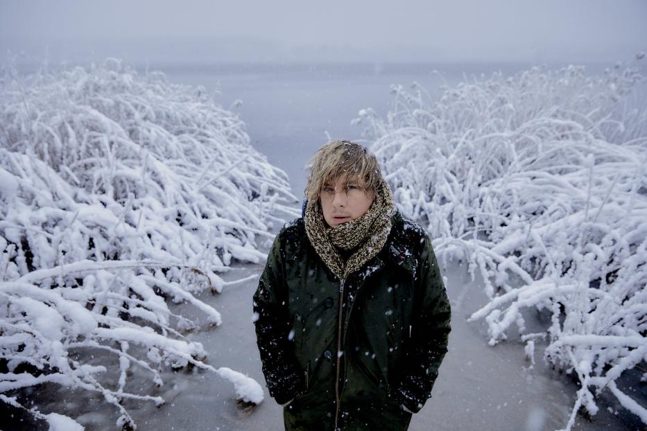 Ein Mann mit Schal und Jacke steht im Schnee zwischen schneebedeckten Pflanzen.