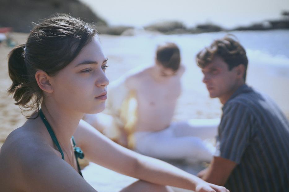 Eine junge Frau sitzt am Strand, im Hintergrund zwei Männer.