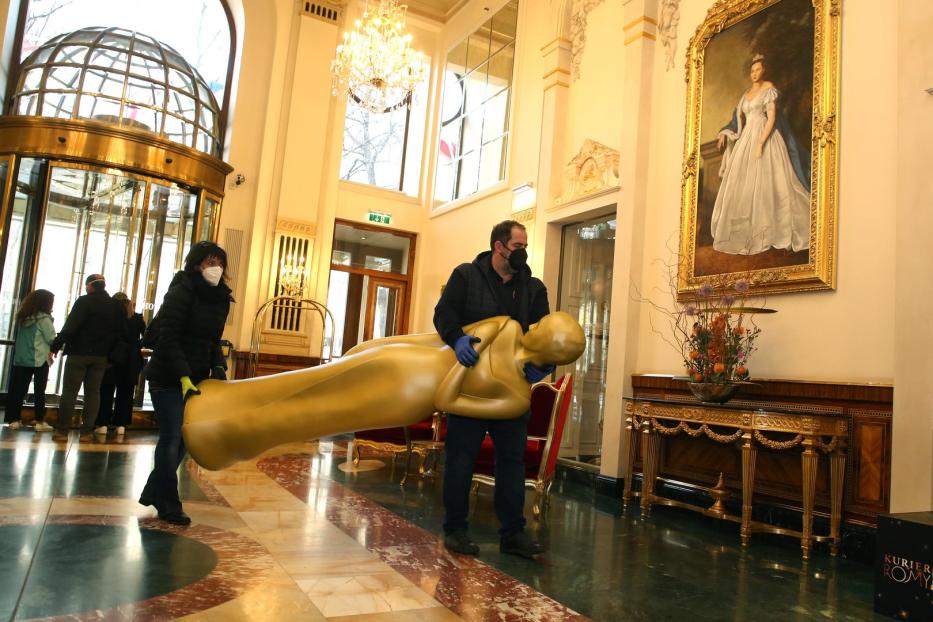 Zwei Personen tragen eine übergroße goldene Oscar-Statue in einem eleganten Gebäude.