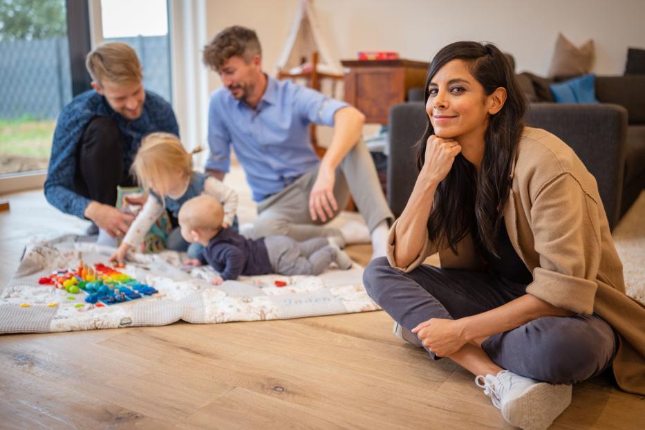 Eine Frau sitzt auf dem Boden, während zwei Männer mit Kindern auf einer Decke spielen.
