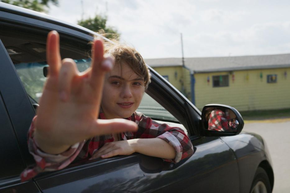 Eine junge Frau lehnt aus einem Auto und formt mit der Hand eine Geste.