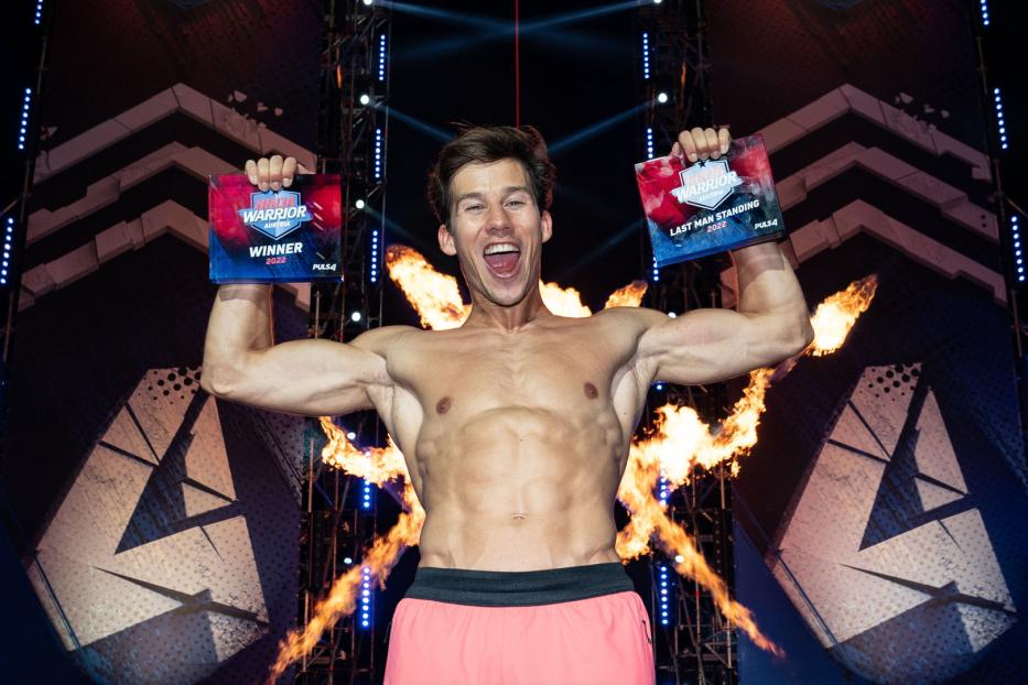 Ein muskulöser Mann jubelt mit zwei „Ninja Warrior Austria“-Trophäen in der Hand vor einer Feuerwand.