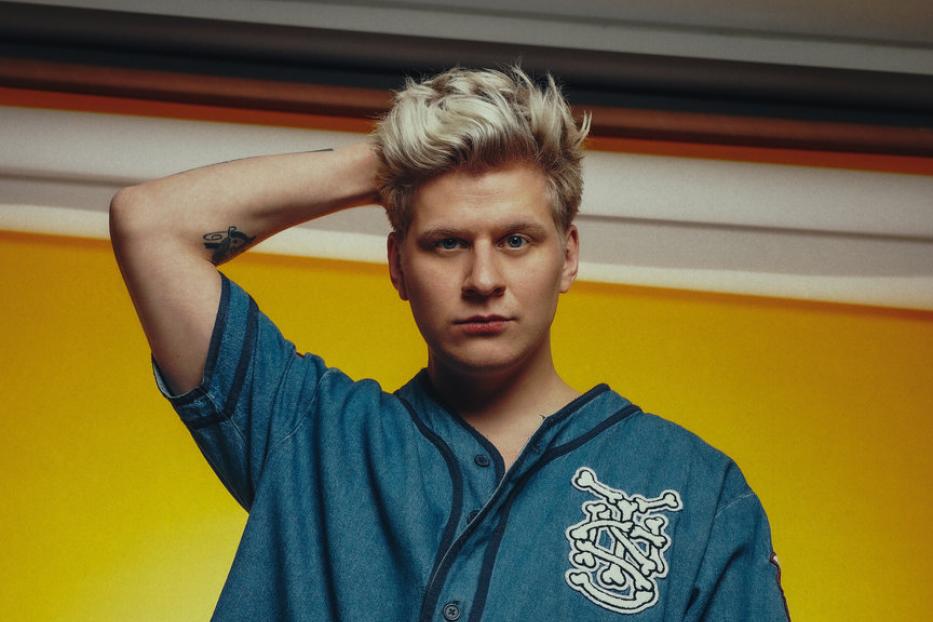 Ein junger Mann mit blonden Haaren posiert vor einem gelben Hintergrund.