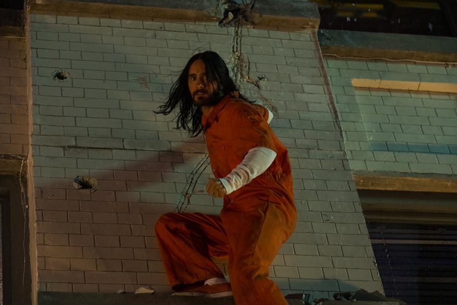 Jared Leto in oranger Gefängniskleidung klettert an einer beschädigten Ziegelmauer hoch.