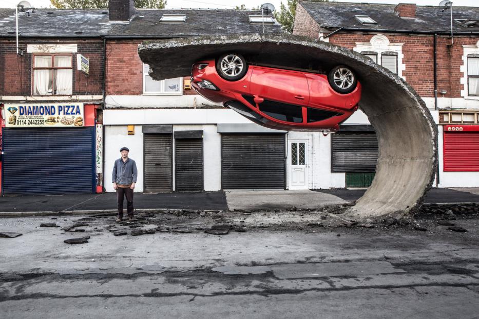 Ein rotes Auto steckt in einem gebogenen Betonstück vor einer Ladenzeile, ein Mann steht davor.