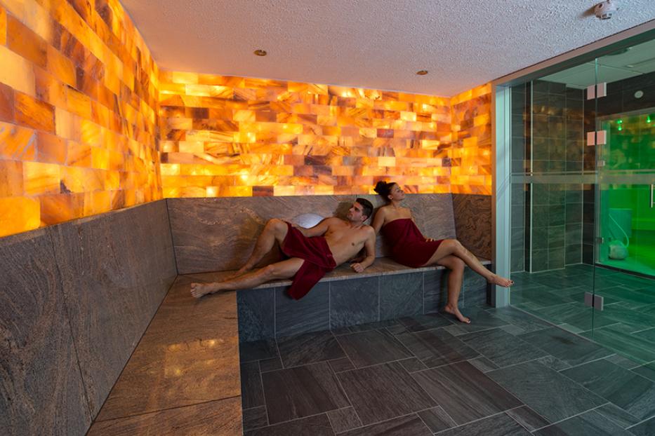 Ein Mann und eine Frau entspannen in Handtücher gehüllt in einer Sauna mit Salzwand.