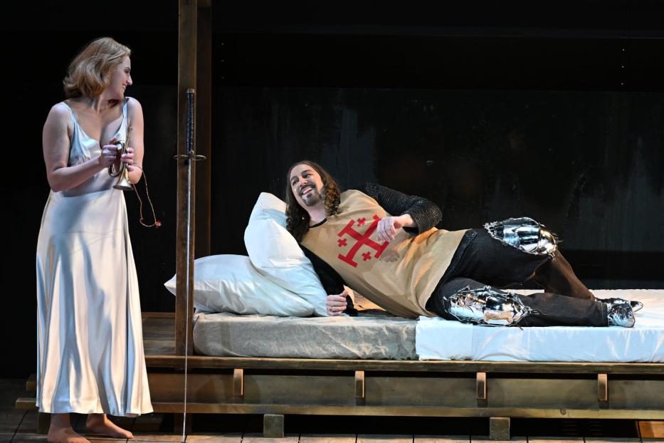 Eine Szene aus einem Theaterstück mit einer Frau mit Trompete und einem Ritter im Bett.