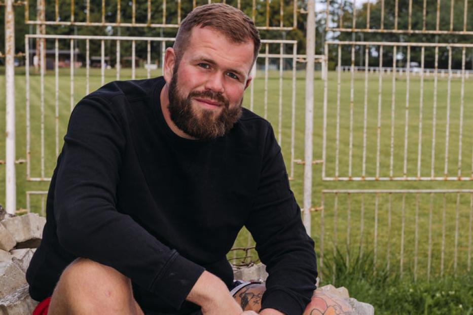 Ein bärtiger Mann mit Tätowierungen sitzt vor einem Fußballplatz auf einem Steinhaufen.