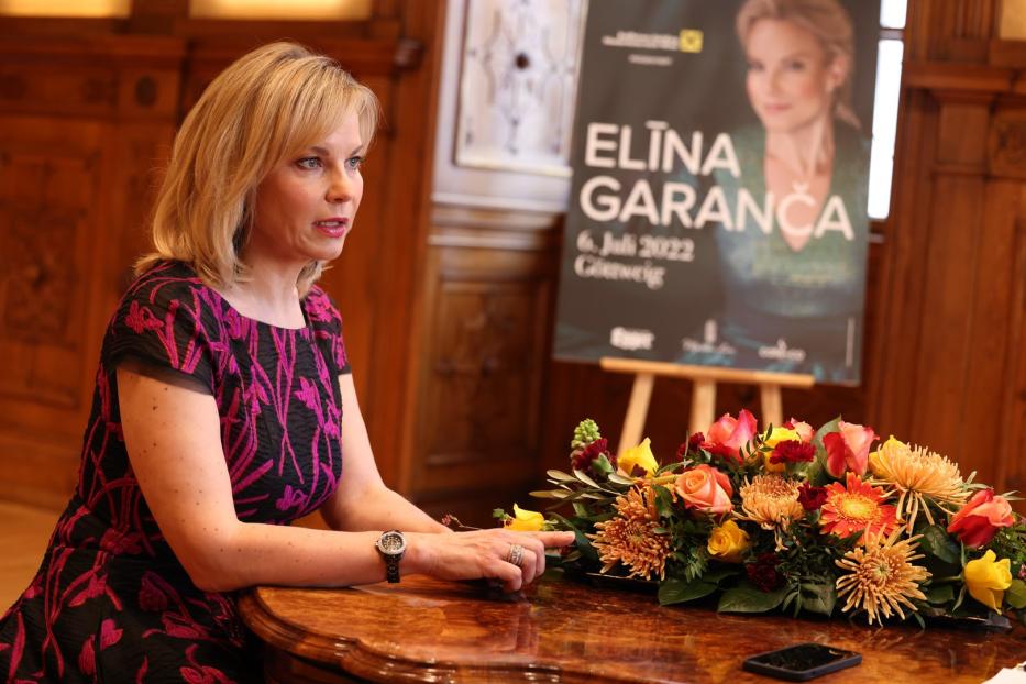 Eine Frau sitzt an einem Tisch mit Blumen; im Hintergrund ein Plakat von Elīna Garanča.