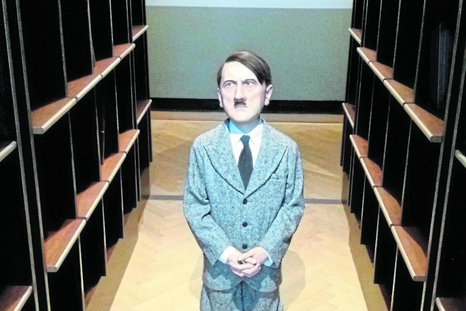 Eine Wachsfigur von Adolf Hitler kniet zwischen leeren Bücherregalen.