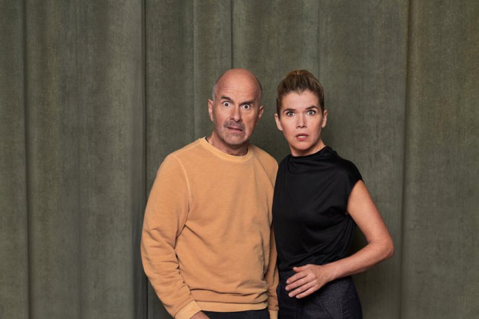 Ein Mann und eine Frau stehen vor einem grünen Vorhang und schauen überrascht in die Kamera.
