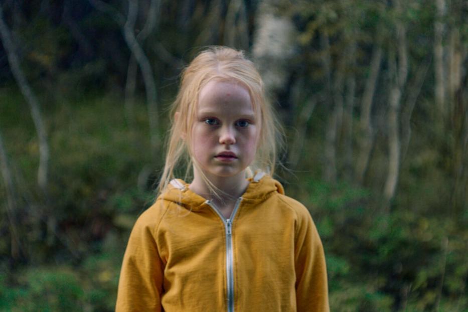 Ein junges Mädchen mit blonden Haaren und gelbem Kapuzenpullover steht vor einem Wald.