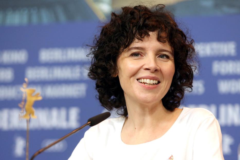 Eine lächelnde Frau mit lockigem Haar vor einem Hintergrund der Berlinale.