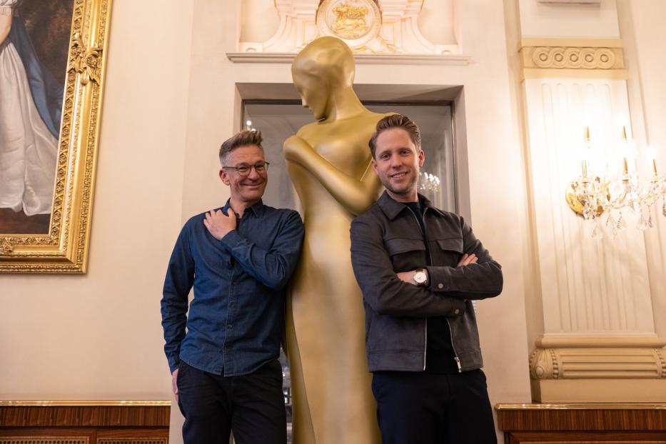 Zwei Männer posieren neben einer goldenen Statue in einem eleganten Raum.
