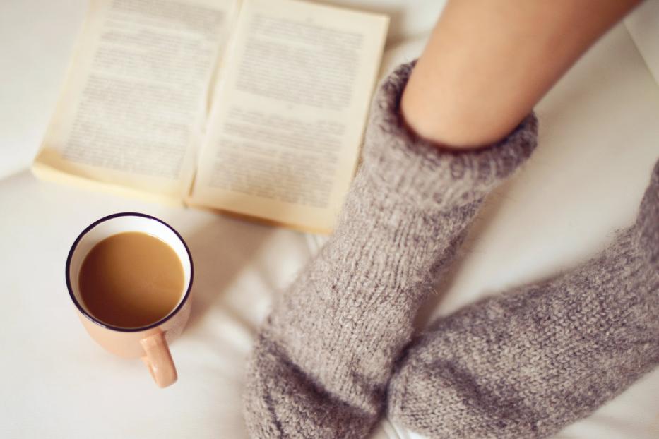 Ein Paar Wollsocken, eine Tasse Kaffee und ein offenes Buch auf einem Sofa.