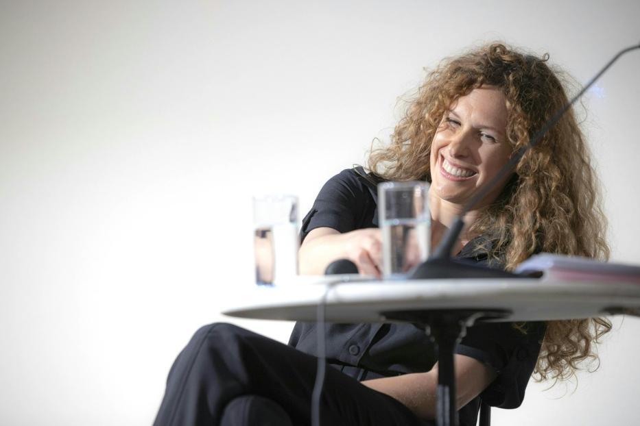 Eine Frau mit lockigem Haar lächelt an einem Tisch mit Mikrofon und Wassergläsern.