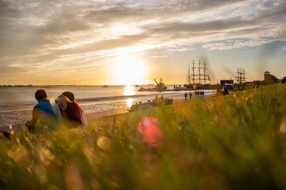 Menschen sitzen am Ufer und beobachten den Sonnenuntergang über dem Meer mit Segelschiffen.