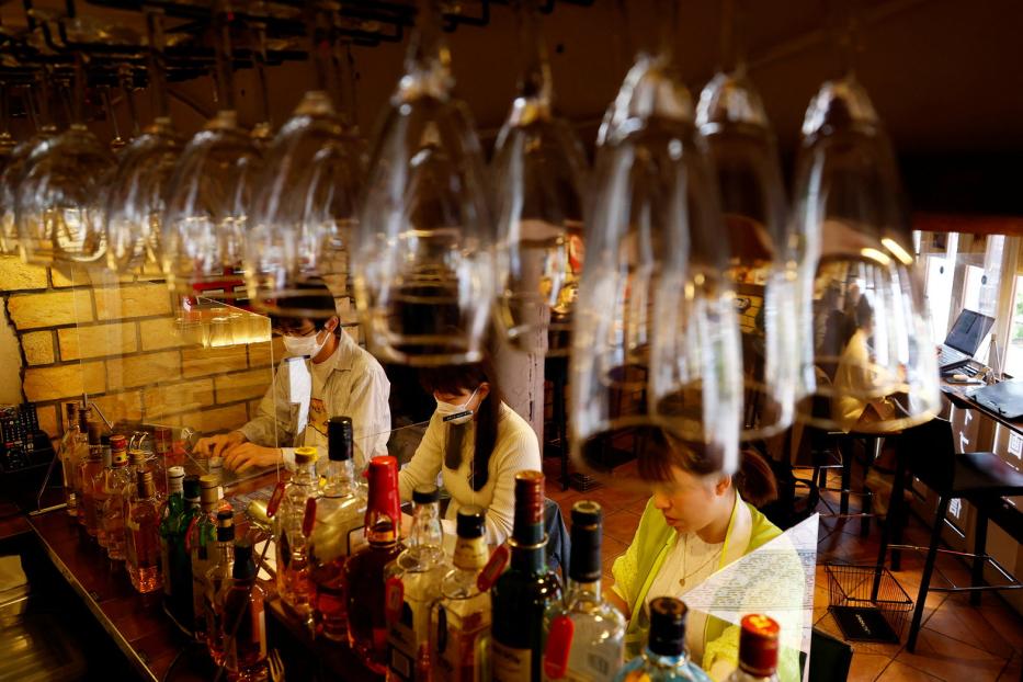 Drei Personen mit Masken arbeiten hinter einer Bar mit vielen Flaschen und Gläsern.