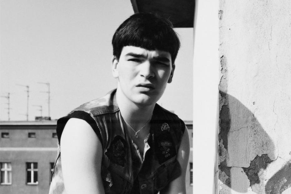 Ein junger Mann mit Bowlcutschnitt und Jeansjacke sitzt vor einem Gebäude.