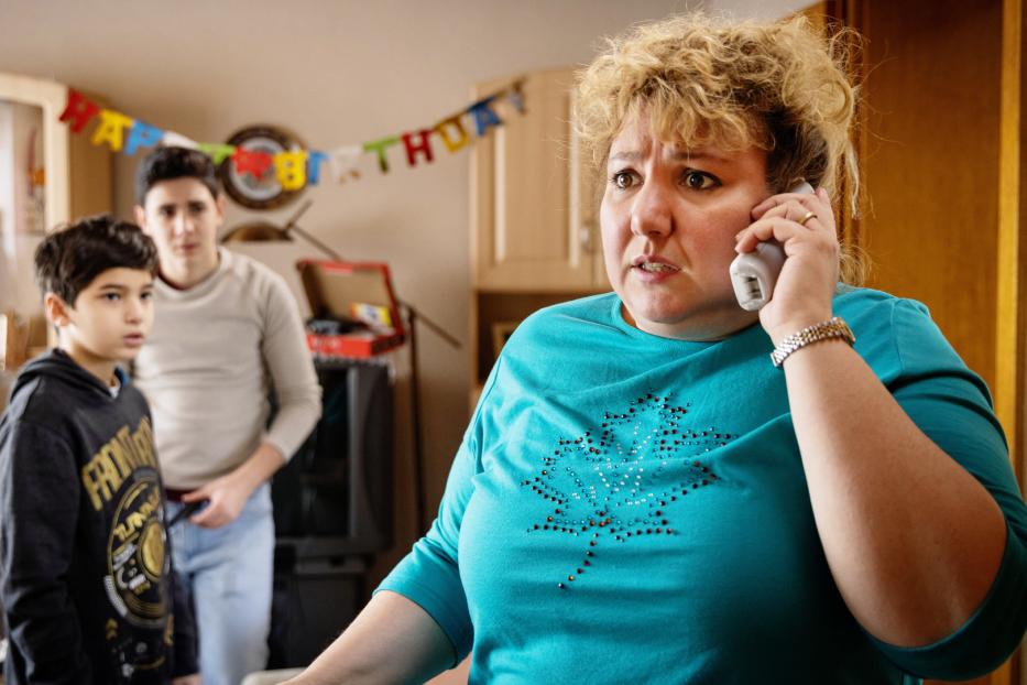 Eine besorgte Frau telefoniert, während zwei Jungen im Hintergrund stehen; eine Geburtstagsgirlande ist zu sehen.