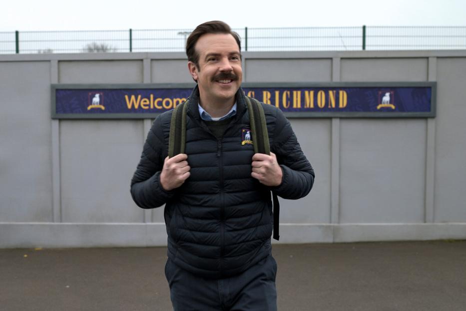 Ted Lasso mit Rucksack vor einem „Welcome to Richmond“-Schild.