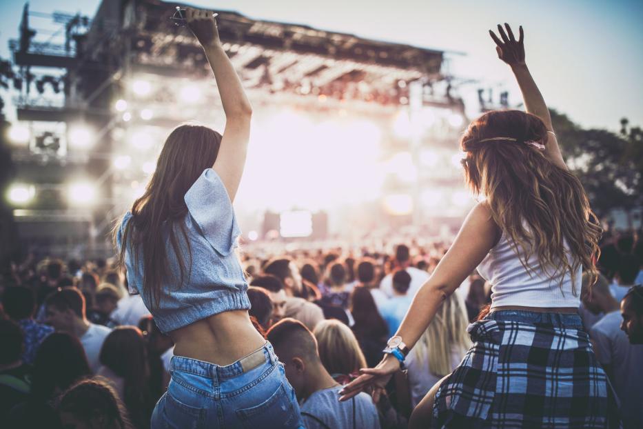 Zwei junge Frauen sitzen auf den Schultern von Freunden bei einem Konzert und jubeln.