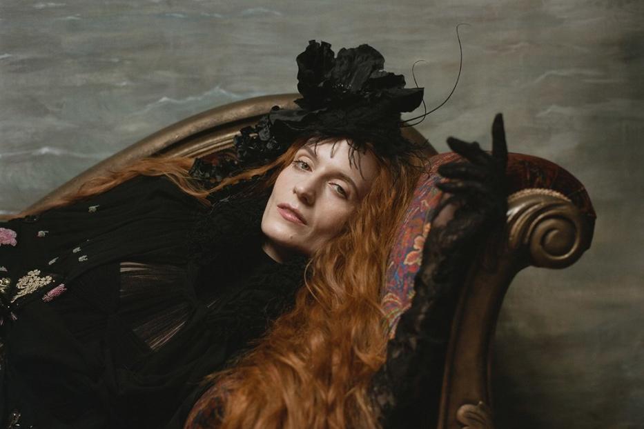 Florence Welch liegt in einem schwarzen Kleid und mit Hut auf einer verzierten Couch.