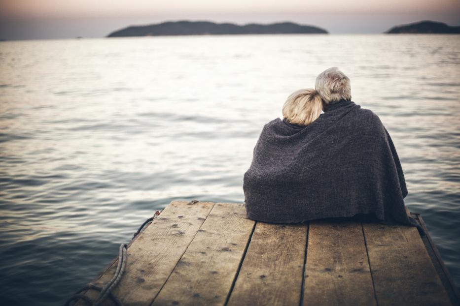 Ein älteres Paar sitzt gemeinsam unter einer Decke auf einem Holzsteg am Meer und blickt auf das Wasser.