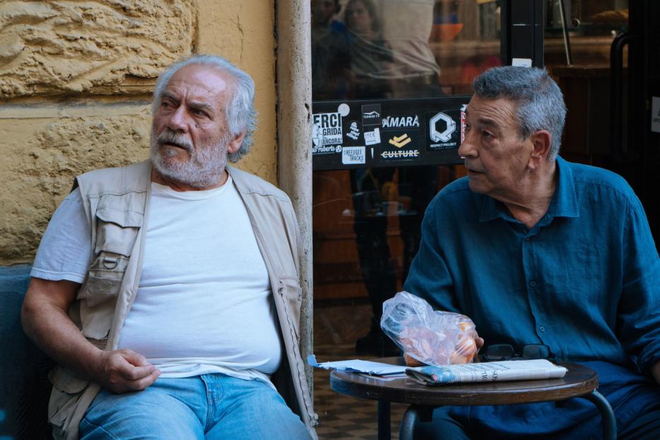 Zwei ältere Männer sitzen an einem Tisch vor einem Café und unterhalten sich.