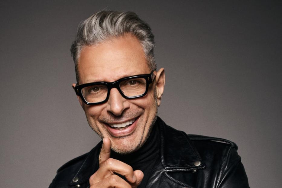 Jeff Goldblum lächelt in schwarzer Lederjacke und Brille, den Finger an die Wange gelegt.