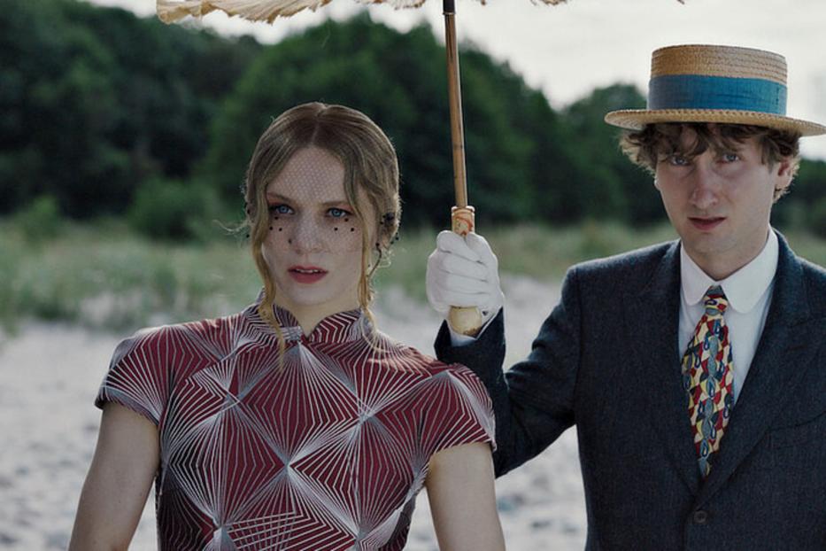 Eine Frau im Retro-Kleid und ein Mann mit Strohhut stehen am Strand.