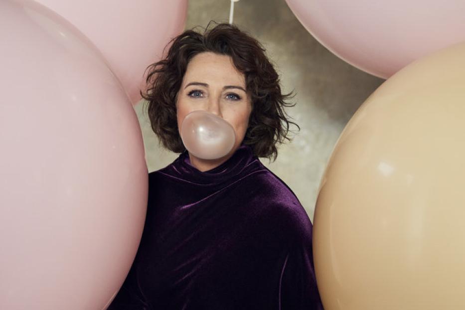 Eine Frau mit dunklen Haaren macht eine Kaugummiblase, umgeben von pastellfarbenen Ballons.