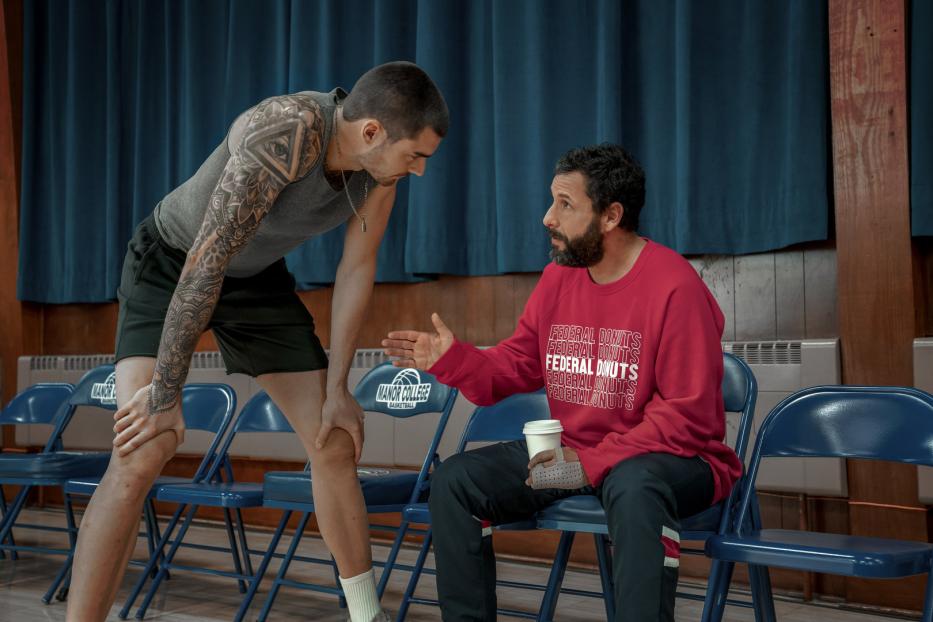 Adam Sandler sitzt mit einem Kaffee in der Hand und spricht mit einem tätowierten Mann.