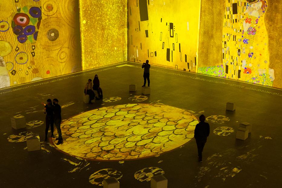Besucher in einer Ausstellung, in der Werke von Gustav Klimt auf Wände und Boden projiziert werden.