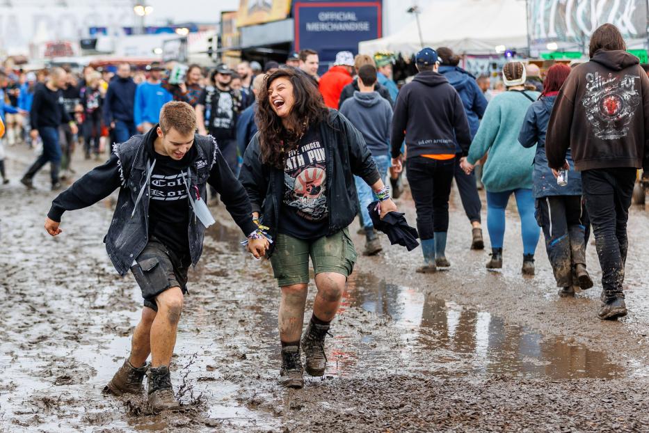 Zwei lachende Personen gehen Hand in Hand durch den Schlamm auf einem Festival.