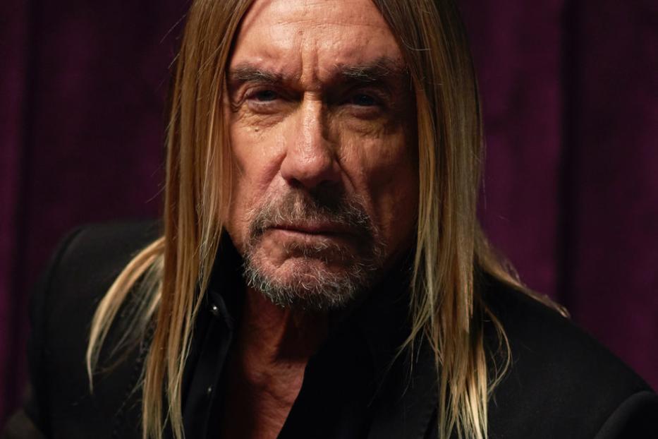 Porträt von Iggy Pop mit langem, blonden Haar und schwarzem Anzug vor einem violetten Hintergrund.