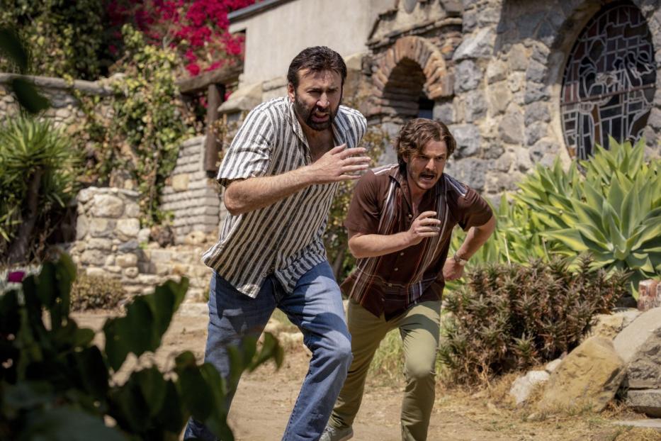 Nicolas Cage und Pedro Pascal rennen in „Massive Talent“ nebeneinander durch eine üppige Landschaft.