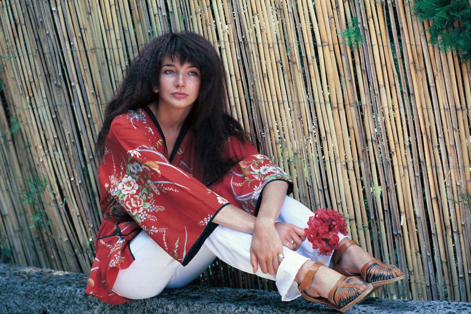 Kate Bush sitzt vor einem Bambushintergrund und hält rote Blumen.