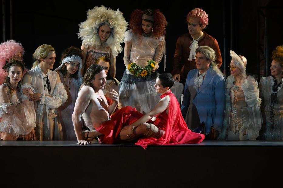 Eine Theaterszene mit einem Mann ohne Hemd und einer Frau in einem roten Kleid, umgeben von Zuschauern in Kostümen.