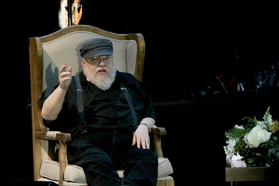 George R.R. Martin sitzt in einem Sessel und gestikuliert während einer Veranstaltung.