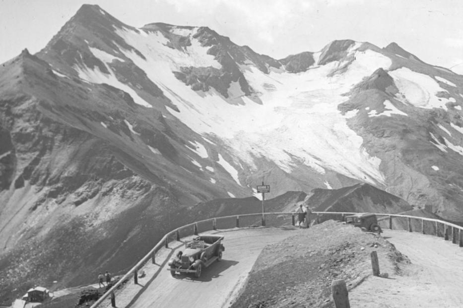 Ein Auto fährt auf einer kurvenreichen Straße vor einer Bergkulisse mit Schnee.