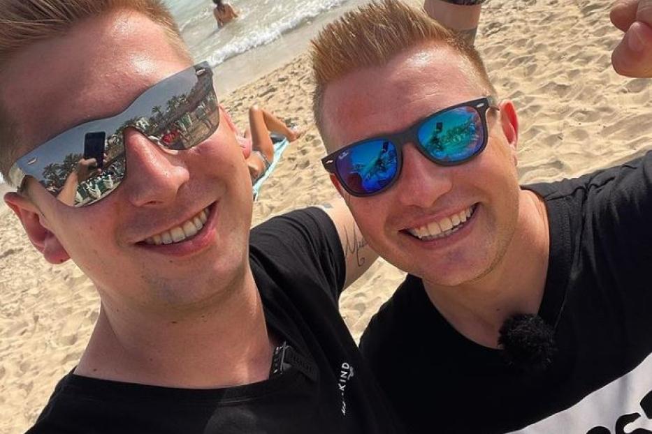 Zwei Männer mit Sonnenbrillen posieren am Strand vor dem Meer.