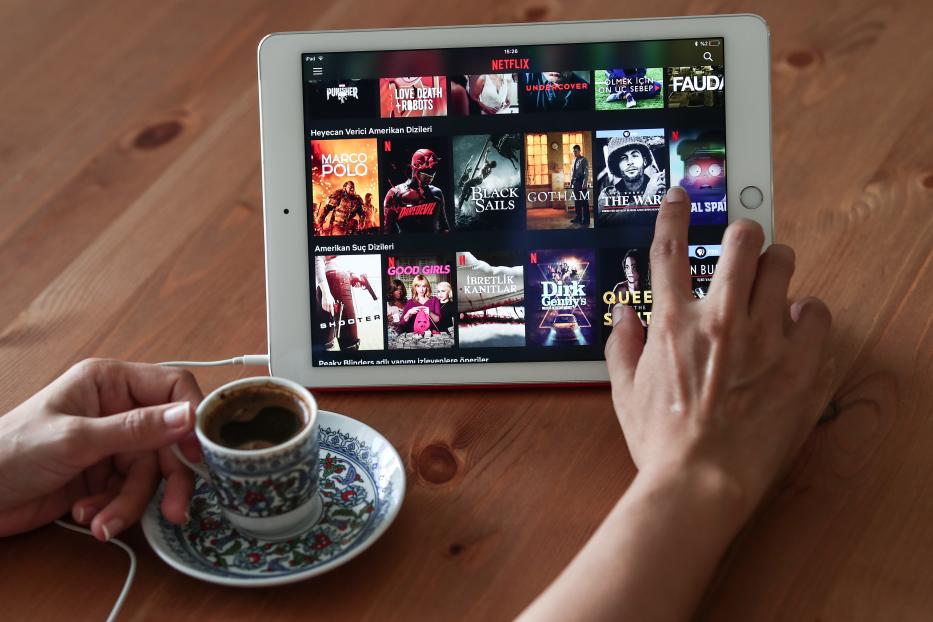 Eine Hand tippt auf einem iPad mit der Netflix-App, daneben eine Tasse türkischer Kaffee.