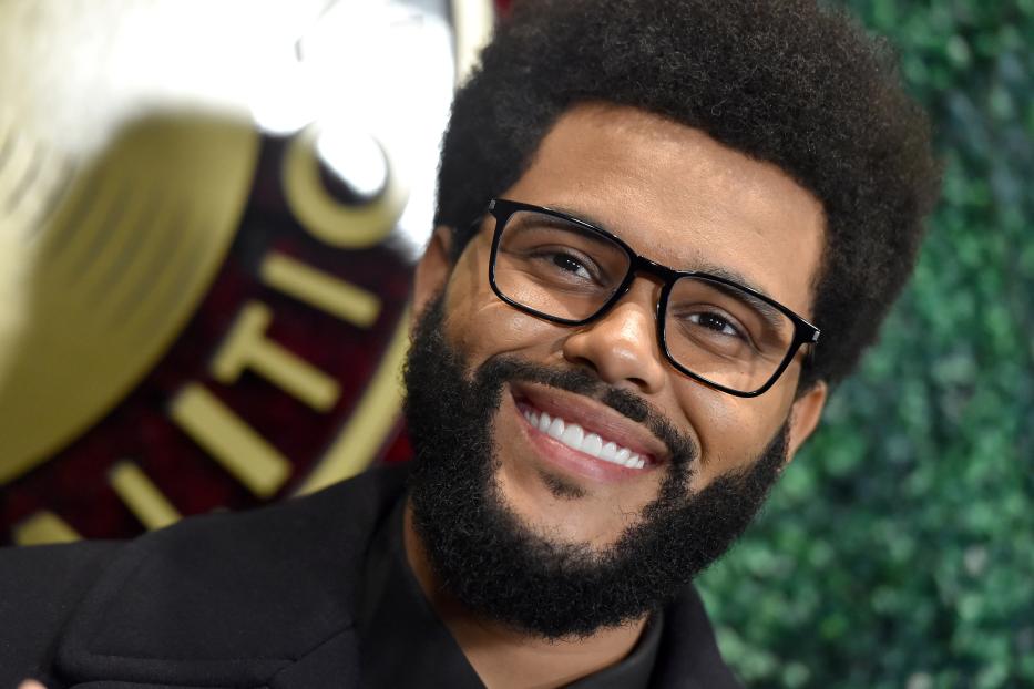 Der lächelnde Sänger The Weeknd mit Brille und Bart vor grünem Hintergrund.