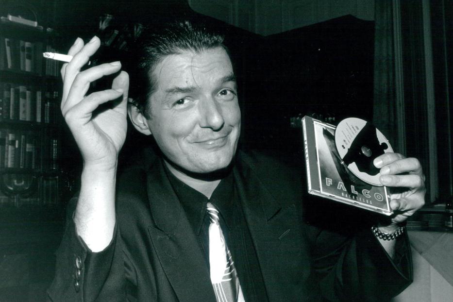 Falco hält eine Zigarette und sein Album „Nachtflug“ in der Hand.